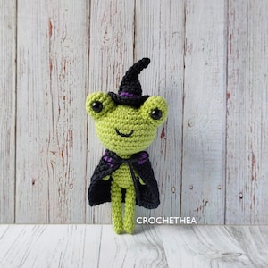 Może przedstawiać: Zielona dziana żaba w czarnym kapeluszu czarownicy z fioletowym wykończeniem i czarną peleryną. Żaba stoi na białym drewnianym podłożu.