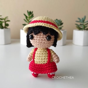 May include: A crocheted doll wearing a yellow dress with red straps, a red and yellow hat, and black hair. The doll is standing on a white surface.