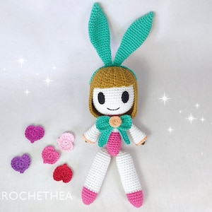 May include: A crocheted doll with bunny ears, a pink bow, and white and pink clothing. The doll is surrounded by small crocheted hearts in various shades of pink.