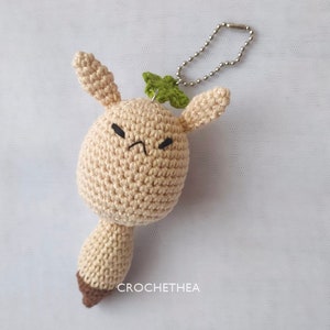 May include: A crocheted beige amigurumi keychain with a green leaf on top. The amigurumi has a brown base and a grumpy face with black eyes.