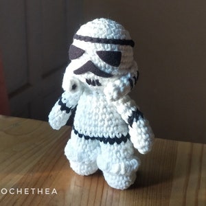 以下が含まれることがあります： 黒のディテールが付いた白い編みぐるみストームトルーパーのフィギュア。フィギュアは木製の表面に立っています。