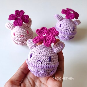May include: Three crocheted plush toys shaped like a round creature with a flower on top. The creatures are purple and pink with embroidered eyes and a purple mouth. The flowers are pink.