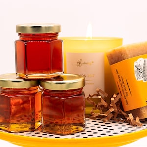 May include: Three small glass jars filled with honey, a yellow candle with the word "thriving" on the front, and a bar of honey soap with the text "Honey Soap, Net Wt 1.5 oz" on the label. The items are arranged on a yellow and black polka dot plate.