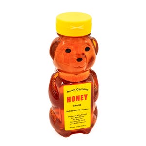 May include: A clear plastic bear-shaped bottle filled with honey. The bottle has a yellow lid and a label that reads "South Carolina Honey Brand Bell Honey Company Producers & Packers of Pure Raw Honey Box 339 Rembert, SC 29128 Net Wt. 12 Oz (340 g)"
