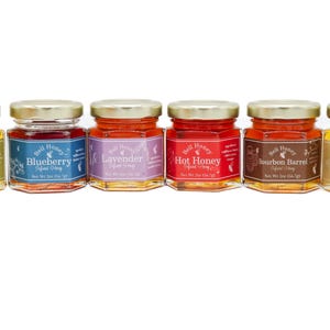 May include: Six small glass jars of infused honey with gold lids are displayed. Flavors include Garlic, Blueberry, Lavender, Hot Honey, Bourbon Barrel, and Vanilla. Each jar has a label with the honey's name and the brand "Bell Honey".