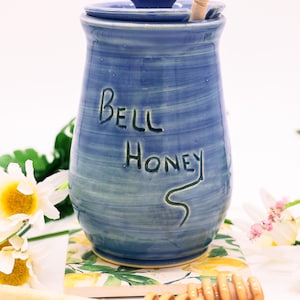 May include: Blue ceramic honey pot with a wooden dipper and the words "Bell Honey" written on the side. The pot is sitting on a lemon-patterned surface with a wooden honey dipper and honey.