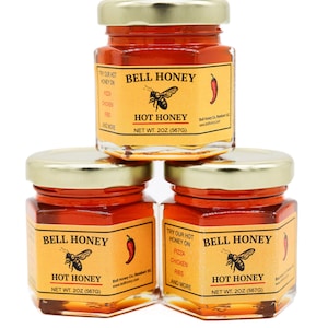 May include: Three glass jars of Bell Honey Hot Honey, each with a yellow label featuring a bee and a red chili pepper. The label reads "Try our hot honey on pizza, chicken, ribs, and more". Each jar contains 2 ounces (567 grams) of honey.