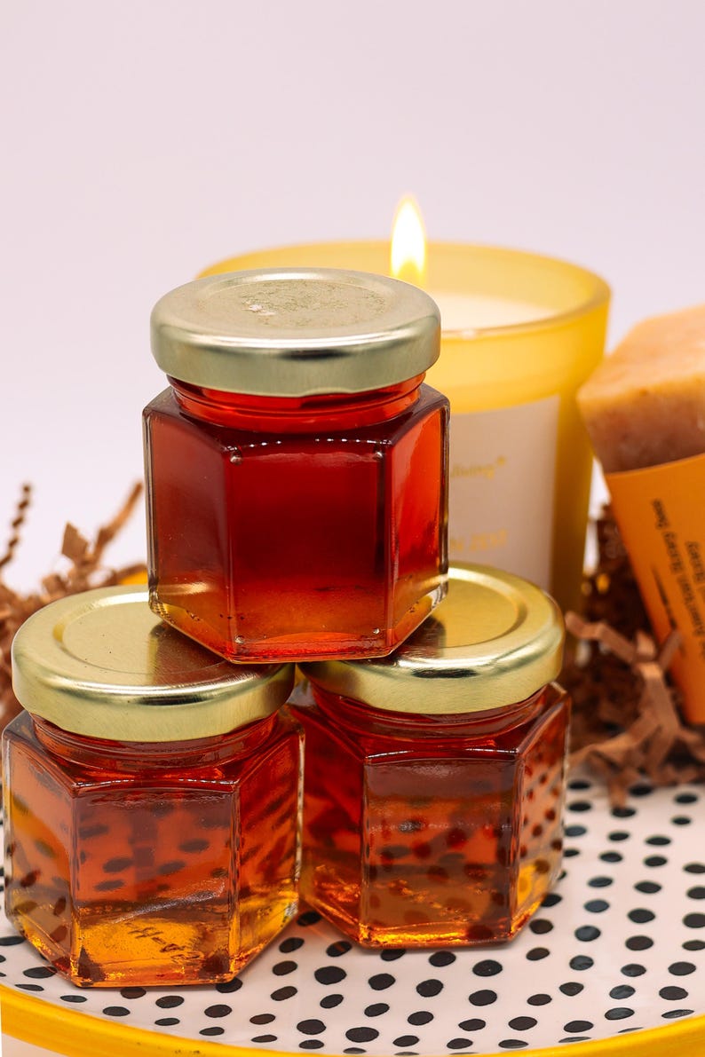 May include: Three small glass jars filled with honey, stacked on top of each other. The jars have gold lids and are on a black and white polka dot surface.