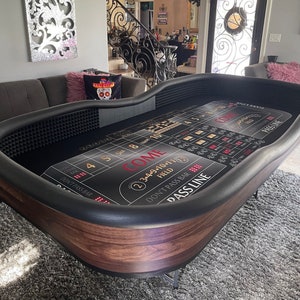 May include: A black felt craps table with a wooden frame. The table has a green felt layout with white and gold markings. The table has a pass line, don't pass bar, come, and don't come bets. The table also has a field bet and a 2/3/4/9/10/11/12 bet.