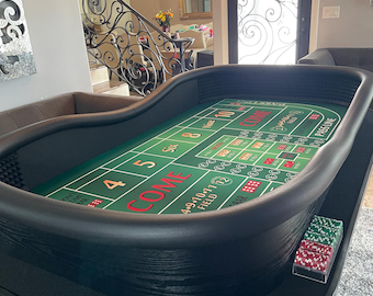 Può includere: Un tavolo da craps con un panno verde caratterizzato da scritte e marcature bianche, rosse e gialle. Il tavolo presenta un bordo imbottito nero e una base in legno nero. È presente un supporto in plastica trasparente con fiches.