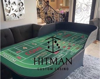 Può includere: Un tavolo da craps da casinò personalizzato con una superficie in feltro verde e finiture nere. Il tavolo presenta segni per varie scommesse, tra cui "PASS LINE" e "COME". Il tavolo ha il testo "HITMAN CUSTOM CASINO" stampato.