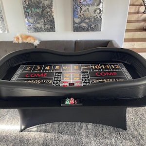 May include: A black craps table with a felt playing surface, featuring numbers, betting areas, and the word "COME." The table has a curved design and is supported by a black base. A few casino chips are visible on the table.