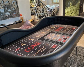 Può includere: Un tavolo da craps nero con una superficie di gioco stampata con le parole "COME" e "PASS BAR". Il tavolo ha un design curvo ed è sostenuto da gambe in metallo nero. Lo sfondo mostra un salotto.