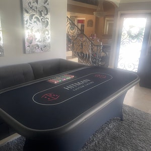 May include: A black felt-covered poker table with a gray felt border. The table has a red dice graphic and the text "Hitman Custom Casino" printed on the surface. The table is set up in a home living room with a gray rug.