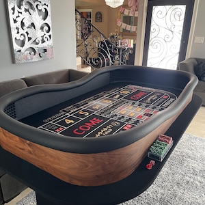 May include: A casino-style craps table with a dark wood finish and black felt playing surface. The table features printed game layout details, including "COME" and number indicators. Dice and chips are visible on the table's edge. The table is covered with a black fabric.