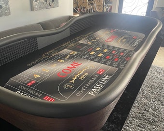 Può includere: Un tavolo da craps rivestito in feltro nero con un bordo in legno marrone. Il tavolo ha un layout in feltro verde con segni bianchi e dorati. Il tavolo ha aree di pass line, don't pass bar, come e don't come. Ci sono due gettoni rossi e verdi sul tavolo.