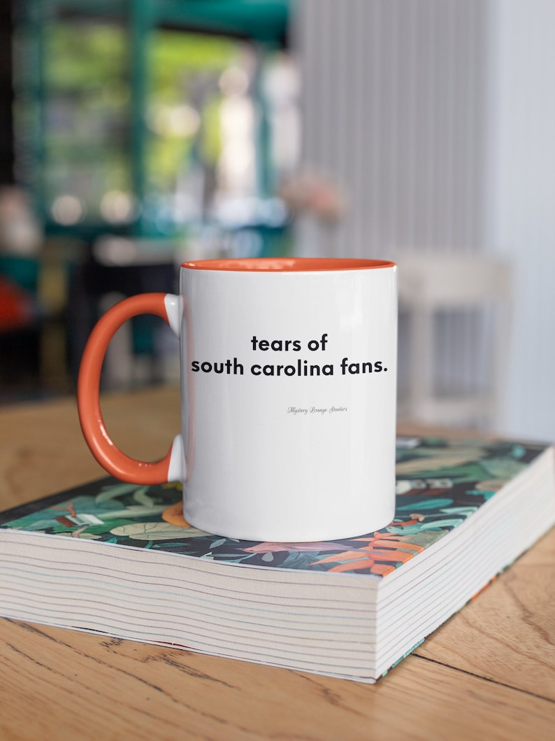Peut inclure: Une tasse en c&eacute;ramique blanche avec un bord et une poign&eacute;e orange. La tasse porte l'inscription "tears of south carolina fans.".