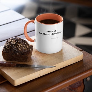Peut inclure: Une tasse &agrave; caf&eacute; blanche avec un bord et une poign&eacute;e orange. La tasse porte une inscription noire qui dit "tears of south carolina fans." La tasse est pos&eacute;e sur une planche &agrave; d&eacute;couper en bois avec un muffin brun fonc&eacute; &agrave; c&ocirc;t&eacute;.