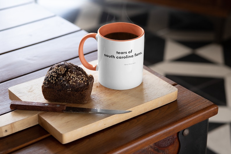 Peut inclure: Une tasse &agrave; caf&eacute; blanche avec un bord et une poign&eacute;e orange. La tasse porte une inscription noire qui dit "tears of south carolina fans." La tasse est pos&eacute;e sur une planche &agrave; d&eacute;couper en bois avec un muffin brun fonc&eacute; &agrave; c&ocirc;t&eacute;.