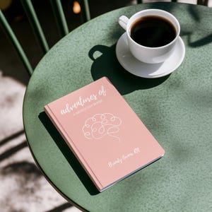 May include: A pink book titled "Adventures of a Neurology Nurse" with a white brain outline and the author's name "Brandy Owens, RN". The book is on a green table with a white coffee cup and saucer.