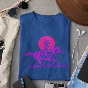 Op de afbeelding: Een koninklijk blauw t-shirt met een roze en paarse afbeelding van een persoon die op een paard rijdt voor een roze zon.