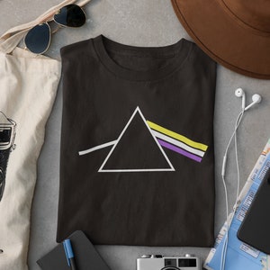 以下が含まれることがあります： 白の三角形とノンバイナリープライドフラッグの色をした虹色のストライプがついた黒いTシャツ。シャツはグレーの表面に、トートバッグ、サングラス、カメラ、ノート、ペン、ヘッドホンなどのアイテムと一緒に置かれています。