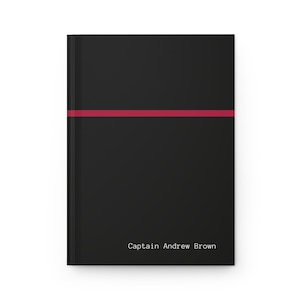 May include: A black journal with a horizontal red stripe across the center. The text "Captain Andrew Brown" is printed in white at the bottom of the journal. The journal has a clean, minimalist design.