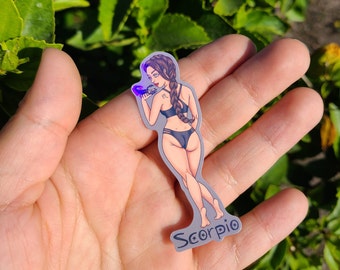 Könnte beinhalten: Ein Aufkleber mit einer Frau mit langen dunklen Haaren, die einen schwarzen Bikini trägt und einen lila Skorpion hält. Der Aufkleber trägt den Schriftzug "Scorpio".