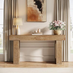 May include: A light brown wooden console table with a thick rectangular top and two sturdy vertical legs. The table surface holds a beige lamp with a textured base, two stacked books, two small candles, and a woven pot containing pink flowers.