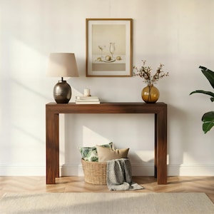 May include: A dark wood console table featuring a table lamp with a beige shade, a framed still life print, a small candle, and a glass vase with branches. A woven basket containing two patterned pillows and a gray throw blanket sits underneath.
