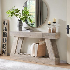 May include: A light wood console table with a rectangular top, two drawers, and a slatted lower shelf. The table features angled side supports. A tall gray textured vase with green foliage, two candle holders, and a woven basket with a white throw.
