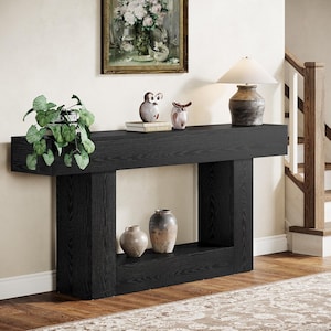 May include: A black wooden console table with a thick rectangular top and two sturdy vertical supports. The table features a lower shelf holding two ceramic vases, while the top surface displays a green potted plant, two owl figurines, and a lamp.
