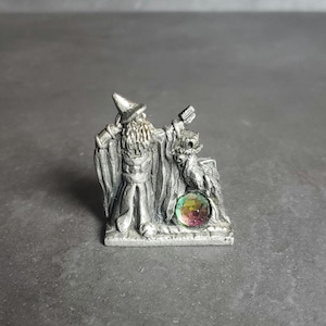 May include: A pewter figurine of a wizard with a long beard and pointed hat, holding a staff and a small dragon. The wizard is standing on a base with a green and purple gemstone.