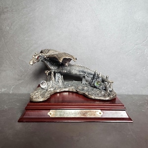 May include: A silver figurine of a flying dragon with red eyes, perched on a rocky base with a green gemstone. The dragon is displayed on a wooden base with a plaque that reads "The Flying Dragon".