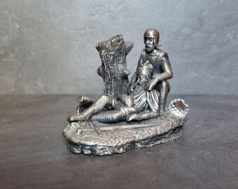 May include: A detailed silver-colored figurine depicting two figures in medieval attire. One figure is seated, supporting a reclining figure. A textured tree trunk and a faceted crystal accent the scene. The figurine is set on a rocky base.
