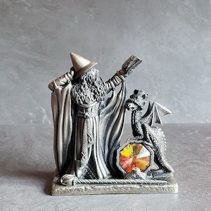 May include: A pewter figurine of a wizard with a long beard and a pointed hat, holding a book and standing next to a small dragon. The wizard and dragon are standing on a base with a large, orange, faceted gemstone.