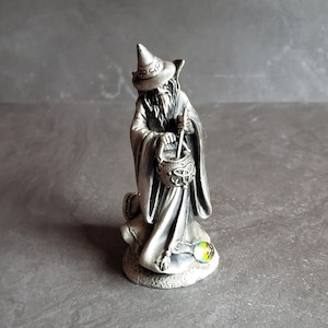May include: A pewter figurine of a wizard stirring a cauldron with a wooden spoon. The wizard is wearing a pointed hat and a long robe. The figurine is standing on a base with a green gemstone.