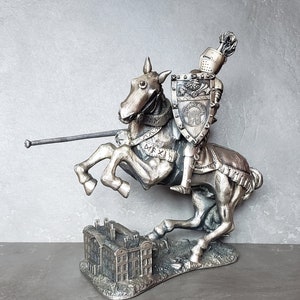 May include: A silver-colored figurine of a knight on horseback, holding a spear and shield. The knight is wearing armor and a helmet with a plume. The horse is rearing up on its hind legs. The figurine is standing on a base that depicts a building.