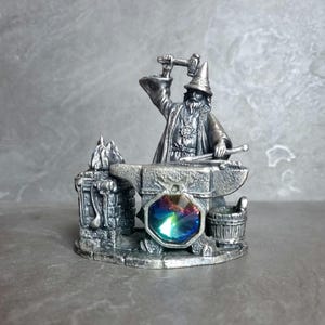 May include: A detailed pewter figurine depicting a wizard blacksmith at work. The wizard, wearing a pointed hat and robe, is hammering on an anvil. A multi-colored, iridescent gemstone is set into the anvil. A small bucket and other tools are also present.