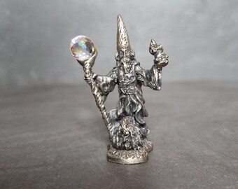 May include: A silver-colored figurine of a wizard, holding a crystal ball in one hand and a small object in the other. The wizard wears a tall, pointed hat and a long robe. The figurine is detailed and stands on a textured base.