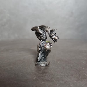 May include: A small, detailed silver-colored dragon figurine perched atop a geometric base. The dragon has outstretched wings and is gazing at a clear, faceted crystal ball. The sculpture is set against a neutral background.