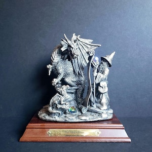 May include: A pewter figurine depicting a wizard with a dragon and a scroll. The wizard is wearing a pointed hat and holding a staff. The dragon is curled around a stone and has a crystal ball in its mouth. The base of the figurine is made of wood and has a plaque that reads "The Taskmaster".