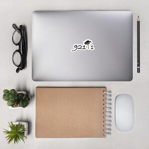 May include: A silver laptop computer with a black and white sticker on the lid. The sticker features a tree silhouette and the text "9211".