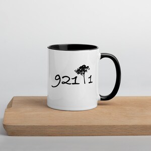 May include: White ceramic mug with a black rim and handle. The mug has a black silhouette of a tree and the text "9211" printed on the front.