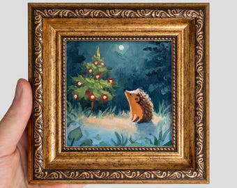 Puede incluir: Una pequeña pintura al óleo en un marco dorado ornamentado de estilo antiguo. La obra muestra a un erizo marrón de pie en un bosque oscuro de tonos azules, cerca de un pequeño árbol decorado con adornos rojos bajo una luna pálida.