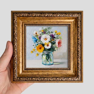 May include: An oil painting of a bouquet of colorful wildflowers in a glass jar, featuring white, yellow, orange, and blue petals with green stems. The artwork is encased in an ornate, textured gold frame with a decorative carved border pattern.
