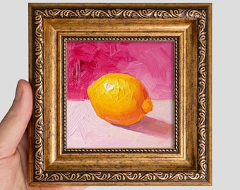 May include: An oil painting of a single yellow lemon centred on a light pink surface against a vibrant magenta background. The artwork is encased in an ornate, antique-style gold frame with intricate embossed detailing along the edges.