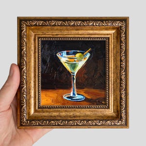May include: An oil painting in a decorative gold frame depicting a martini glass filled with a pale yellow liquid and a single green olive on a toothpick. The background is dark, and the glass sits on a warm, orange-toned surface.