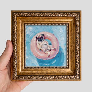 May include: An oil painting of a pug wearing pink sunglasses while floating in a pink inner tube on blue water. The artwork is encased in an ornate, antique-style gold frame with a textured, carved border and a deep, polished finish.