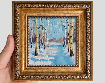 May include: An oil painting of a winter forest scene featuring white birch trees with dark markings set against a light blue sky and snow-covered ground, enclosed in a square, ornate, metallic gold frame with a decorative embossed border.